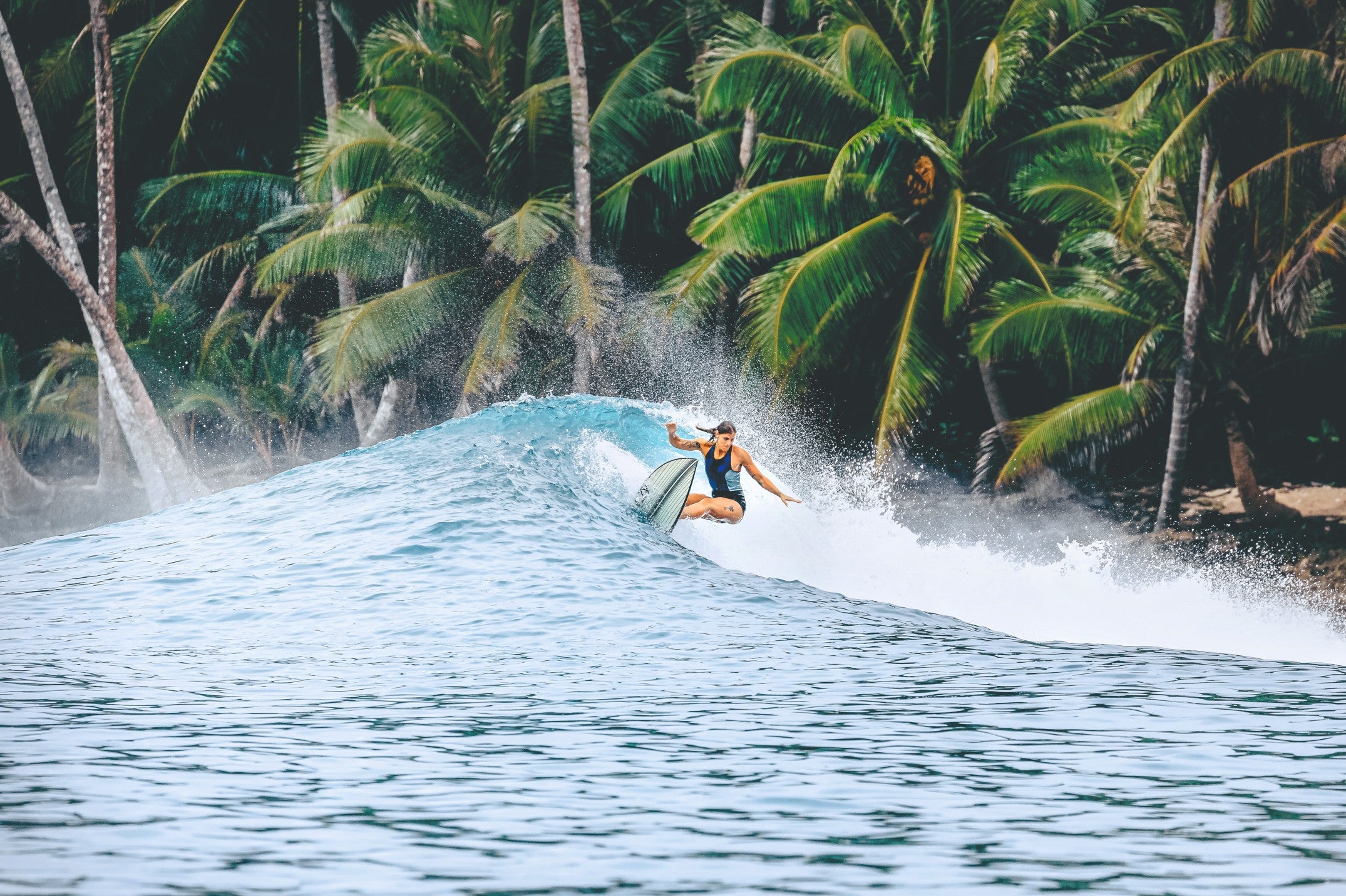 women surfing a wave with a short board. She wears a swimsuit and in the back are palm trees